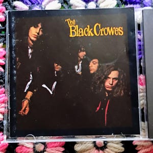 May include: A CD case featuring the band The Black Crowes. The album cover has a dark background with a photo of the band members. The band's name is in yellow text at the top.