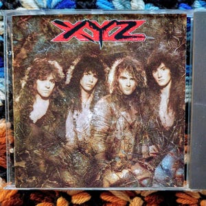 May include: A CD case featuring the band XYZ. The album cover has a textured, sepia-toned background with the band members in the center. The band's name, XYZ, is in red block letters at the top. The band members have long hair and are wearing jackets.