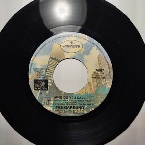 May include: A black vinyl record with a white label. The label has a picture of a city skyline and the text "Who Do You Call" by "The Gap Band". The record is from Mercury Records and is part of the album "The Gap Band II".