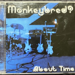 May include: CD case with the band name "Monkeybred?" in white letters. The album cover is a blue-toned image of a band's instruments, including guitars, drums, and amplifiers. The album title "About Time" is at the bottom.