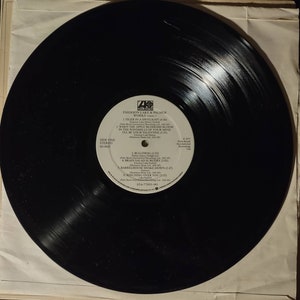 Puede incluir: Disco de vinilo negro con una etiqueta blanca. La etiqueta tiene el texto "Emerson Lake & Palmer Works" y enumera las canciones "Tiger in a Spotlight", "When the Apple Blossoms Bloom in the Windmills of Your Mind", "I'll Be Your Valentine", "Bullfrog", "Brain Salad Surgery", "Barrelhouse Shake Down" y "Watching Over You".