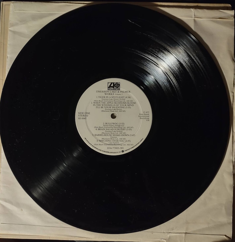 Puede incluir: Disco de vinilo negro con una etiqueta blanca. La etiqueta tiene el texto "Emerson Lake & Palmer Works" y enumera las canciones "Tiger in a Spotlight", "When the Apple Blossoms Bloom in the Windmills of Your Mind", "I'll Be Your Valentine", "Bullfrog", "Brain Salad Surgery", "Barrelhouse Shake Down" y "Watching Over You".