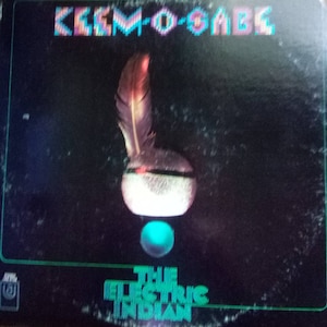 May include: A black vinyl record album cover with the title "The Electric Indian" in green text. The album cover features a colorful feather and a turquoise circle on a black background. The artist's name, "Kemo-Sabe", is printed in colorful block letters at the top of the cover.