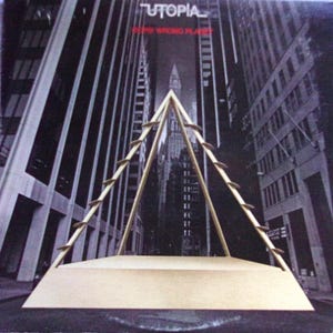 May include: A black and white photo of a city skyline with a gold metal structure in the foreground. The structure is a triangular frame with a flat top. The text "Utopia" and "The Wrong Planet" is visible at the top of the image.