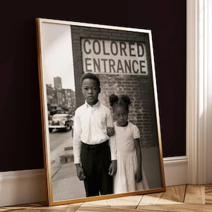 以下が含まれることがあります： 「COLORED ENTRANCE」の標識があるレンガの壁の前に立っている2人の子供が写った、白黒の額入り写真。男の子は白いシャツと黒いズボン、女の子は白いドレスを着ています。フレームは金色です。