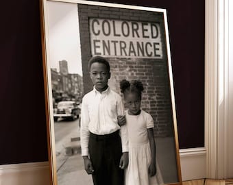 May include: Black and white framed photograph featuring two children standing in front of a brick wall with a sign that reads "COLORED ENTRANCE". The boy wears a white shirt and black pants, and the girl wears a white dress.