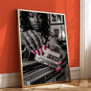 May include: A framed black and white photograph featuring a woman holding a cassette tape labeled "Positive Black Music." The woman's hands, adorned with rings and long pink nails, are the focal point. The cassette tape rests on a vintage boombox.
