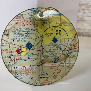 May include: A round, clear ornament featuring a detailed aviation sectional chart. The chart includes airport identifiers, frequencies, and navigational aids. A gold string is attached for hanging. The ornament is a unique gift for pilots or aviation enthusiasts.