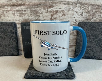 May include: White ceramic mug with a blue rim and handle. The mug has a graphic of a small airplane and the text "FIRST SOLO" in black. Below the airplane is the text "John Smith", "Cessna 172 N12345", "Kansas City, KMKC", and "December 1, 2021".