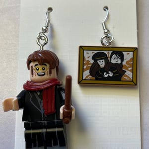 Peut inclure: Une paire de boucles d'oreilles pendantes avec des figurines de style Lego. Une boucle d'oreille représente une figurine avec des cheveux bruns, une écharpe rouge et une baguette. L'autre boucle d'oreille montre une image encadrée de deux personnages.