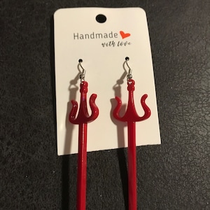 May include: A pair of red, plastic earrings shaped like a trident. The earrings are hanging from a white card with the text "Handmade with love".