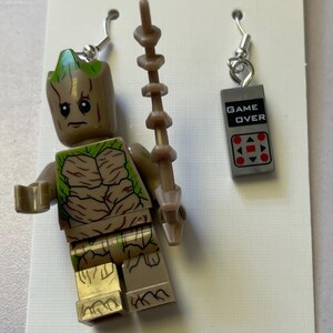 May include: A pair of earrings featuring a Groot figure and a gray game controller charm. The Groot earring is a brown and green Lego figure with a staff. The other earring has the words "GAME OVER" and a red button design.