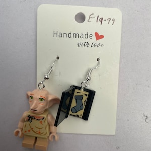 以下が含まれることがあります： レゴ風のフィギュアと靴下のグラフィックが入った黒い本が特徴の手作りのイヤリング。フィギュアはタン色で、緑色の目とベージュの衣装をしています。イヤリングは銀色のフックから吊り下げられており、「Handmade with love」の文字が入った白いカードに付いています。