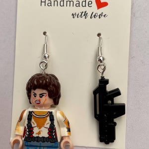 May include: Handmade earrings featuring a Lego figure and a black gun. The Lego figure has brown hair and is wearing a white shirt with orange stains, blue jeans, and red shoes. The other earring is a black gun.