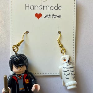 May include: Handmade earrings featuring a Harry Potter Lego figure and a white owl Lego figure. The earrings have gold-colored hooks. The card behind the earrings says "Handmade with love" in black text with a red heart.