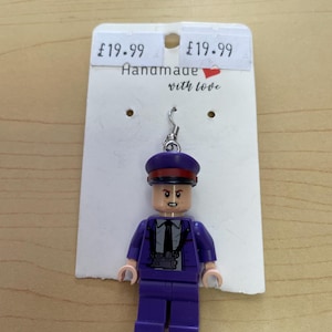 Puede incluir: Un pendiente artesanal estilo Lego con una figura uniformada púrpura con corbata negra y sombrero. El pendiente está sujeto a un gancho plateado. La etiqueta dice "Handmade with love".