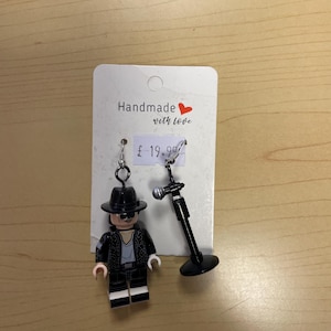 May include: Handmade earrings featuring a Lego figure in a black hat and jacket, and a miniature black microphone. The earrings are attached to a white card with the text "Handmade with love".