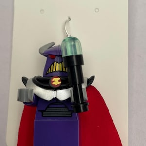 May include: Purple and silver Lego figure with a red cape and a black weapon. The figure has a green light on its head. The figure is hanging from a silver earring hook. The tag says "Handmade with love" and "£19.99".