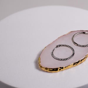 Peut inclure: Deux boucles d'oreilles créoles argentées avec un design torsadé, présentées sur un sous-verre en agate rose et or.