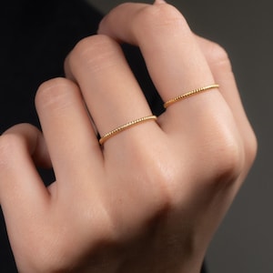 以下が含まれることがあります： 手には、テクスチャ加工されたビーズのデザインが施された、2つの繊細なゴールドリングが着用されています。 リングは細くてミニマルで、エレガントさを添えています。 背景は濃いグレーで、ジュエリーを際立たせています。