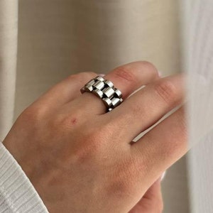 May include: A silver chain link ring with a square design. The ring is worn on a hand with white long-sleeved shirt.