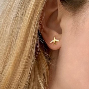 Può includere: Una donna indossa una maglietta bianca con una collana a catena dorata e orecchini a forma di aereo dorati.