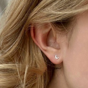 Könnte beinhalten: Nahaufnahme eines Ohrs mit einem kleinen Halbmond-Ohrring. Der Ohrring ist aus Silber und hat ein schlichtes, zartes Design.