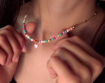Może przedstawiać: Kolorowy naszyjnik choker z koralików z małym różowym serduszkiem. Naszyjnik wykonany jest z małych, okrągłych koralików w różnych odcieniach różu, błękitu, zieleni i bieli. Koraliki są nawleczone na złoty łańcuszek.
