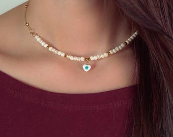 Może przedstawiać: Naszyjnik choker z białych koralików z złotym łańcuszkiem i serduszkowym zawieszką z niebieskim oczkiem.