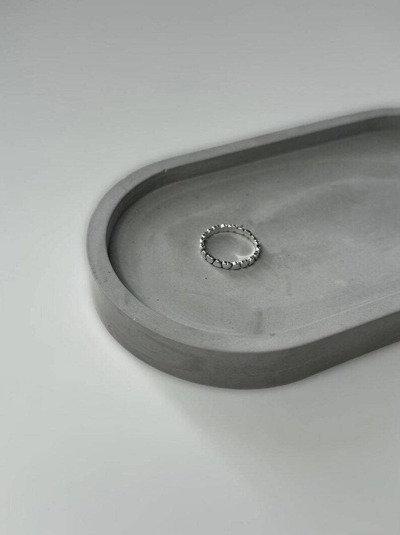 May include: A silver ring with a beaded design sits on a gray concrete dish.