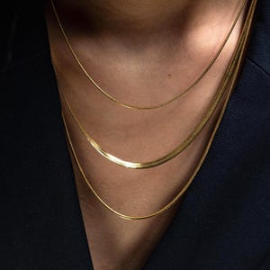 Könnte beinhalten: Drei goldene Ketten, die auf dem Hals einer Person übereinander liegen. Die Ketten haben unterschiedliche Breiten und Stile, wobei die oberste Kette die dünnste und die unterste Kette die dickste ist.