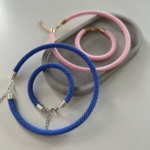 Pu&ograve; includere: Un set di tre collane di perline blu e rosa. Le collane sono fatte di piccole perline e hanno chiusure argentate. Le collane sono disposte su una superficie grigia.