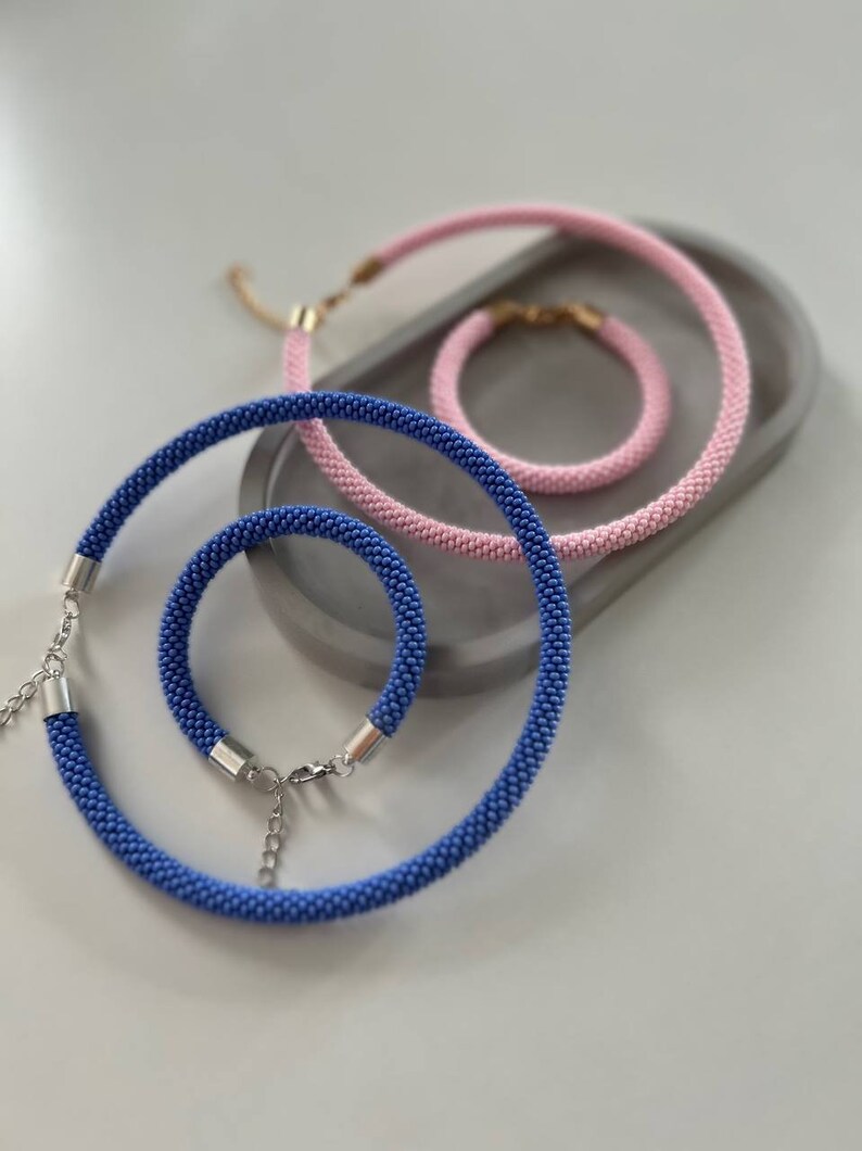 Pu&ograve; includere: Un set di tre collane di perline blu e rosa. Le collane sono fatte di piccole perline e hanno chiusure argentate. Le collane sono disposte su una superficie grigia.