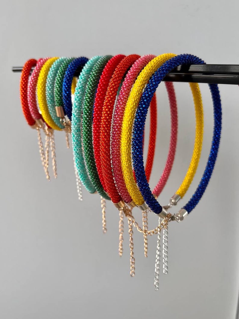 Pu&ograve; includere: Un set di braccialetti colorati con perline e dettagli di catena dorata e argentata. I braccialetti sono disposti a forma di arcobaleno, ogni braccialetto &egrave; di un colore diverso.
