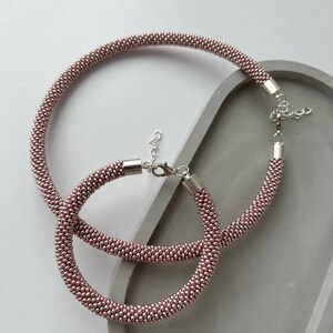 Pu&ograve; includere: Due collane di perline color oro rosa con chiusure argentate. Le collane sono disposte su una superficie grigia.