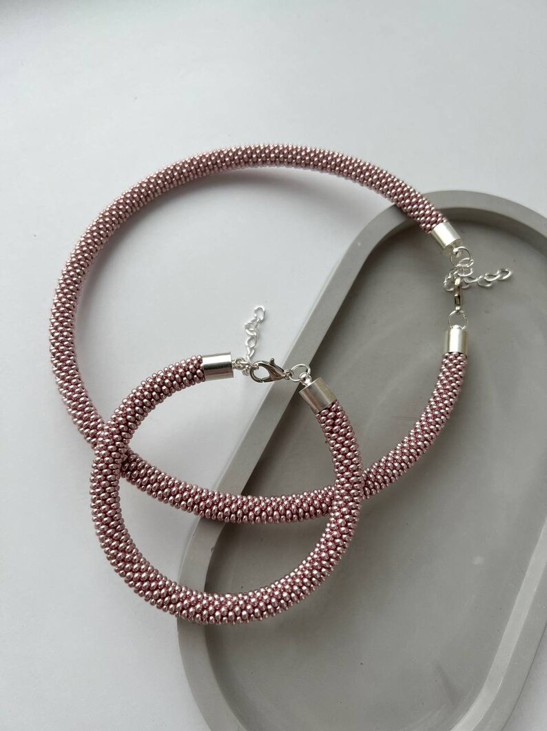 Pu&ograve; includere: Due collane di perline color oro rosa con chiusure argentate. Le collane sono disposte su una superficie grigia.