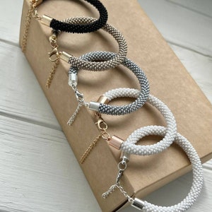 Pu&ograve; includere: Quattro braccialetti di perline in nero, argento, oro e bianco. I braccialetti sono impilati uno sopra l'altro su una scatola marrone. Ogni braccialetto ha una catenina dorata e una chiusura argentata.