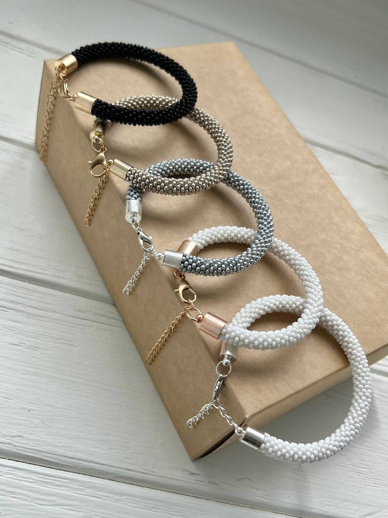 Pu&ograve; includere: Quattro braccialetti di perline in nero, argento, oro e bianco. I braccialetti sono impilati uno sopra l'altro su una scatola marrone. Ogni braccialetto ha una catenina dorata e una chiusura argentata.