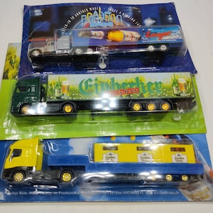 May include: Three toy semi-trucks on their original packaging. The trucks are various colors, including yellow, green, and blue. The trailers feature beer-related graphics and text. The packaging includes the text "CHECK-IN TO ANOTHER WORLD" and "TRATE A BINDING LAS".