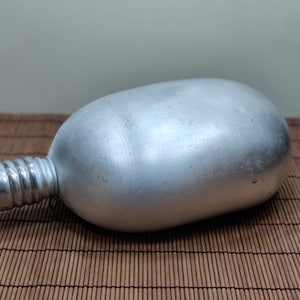 May include: A silver-colored canteen with a screw-on cap. The canteen has a rounded body and a textured neck. The canteen is made of metal and sits on a bamboo mat.