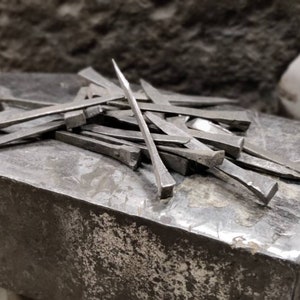 May include: A pile of hand-forged iron nails on a metal anvil. The nails are various sizes and shapes, with some having pointed tips and others having flat heads.