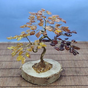 May include: A decorative amber tree sculpture with a twisted gold-colored wire trunk and branches. The leaves are made of amber-colored stones, ranging from light yellow to dark brown. The tree sits on a round wooden base.