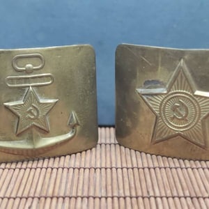 May include: Two gold-colored metal belt buckles. The buckle on the left features an anchor with a red star and a hammer and sickle inside the star. The buckle on the right features a red star with a hammer and sickle inside the star.