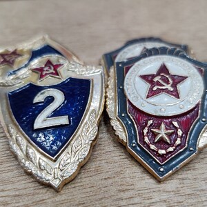 Op de afbeelding: Twee vintage Sovjet-tijdperk badges. Eén heeft een blauw schild met het nummer 2, een rode ster en een hamer en sikkel. De andere heeft een rode ster, hamer en sikkel en tekst in een cirkelvormig ontwerp.