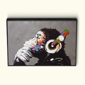 Peut inclure: Une œuvre d'art encadrée représentant un chimpanzé portant un casque audio. Le visage du singe est partiellement peint avec des éclaboussures colorées, et le casque a un design arc-en-ciel. Le fond est gris uni.