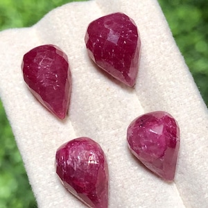 May include: Three faceted, pear-shaped rubies with a deep magenta-red colour. Each gemstone features a polished surface with multiple geometric facets that reflect light, showcasing a translucent quality and a distinct teardrop silhouette.