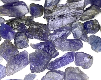 May include: A collection of raw tanzanite gemstones in various shapes and sizes. The stones display shades of deep blue and purple, with some appearing nearly black. The rough, natural texture of the gemstones is visible against a white background.