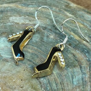 May include: A pair of gold-colored high heel earrings with black accents. The earrings have silver-colored hooks and are adorned with small, clear rhinestones on the heels. The earrings are designed to resemble miniature high-heeled shoes.