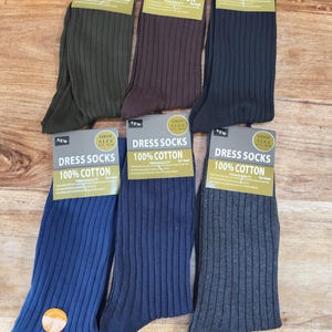 May include: Six pairs of ribbed dress socks made of 100% cotton, displayed in olive green, brown, black, bright blue, navy blue, and charcoal gray. Each pair features a cardboard header tag labeled DRESS SOCKS 100% COTTON and shoe size 6-11.