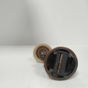 May include: Close-up of a vintage pencil sharpener with a bronze-colored metal base and a clear, cylindrical plastic top. The sharpener has a blade visible inside and is set against a white background.
